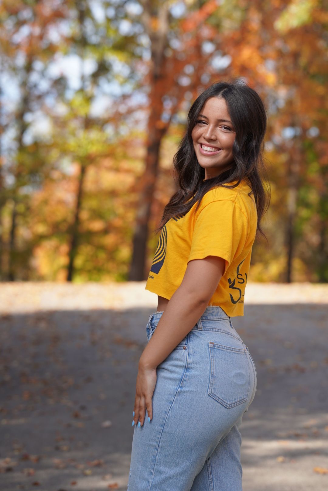 Woman in yellow t-shirt and light denim jeans poses outdoors against autumn foliage background.
