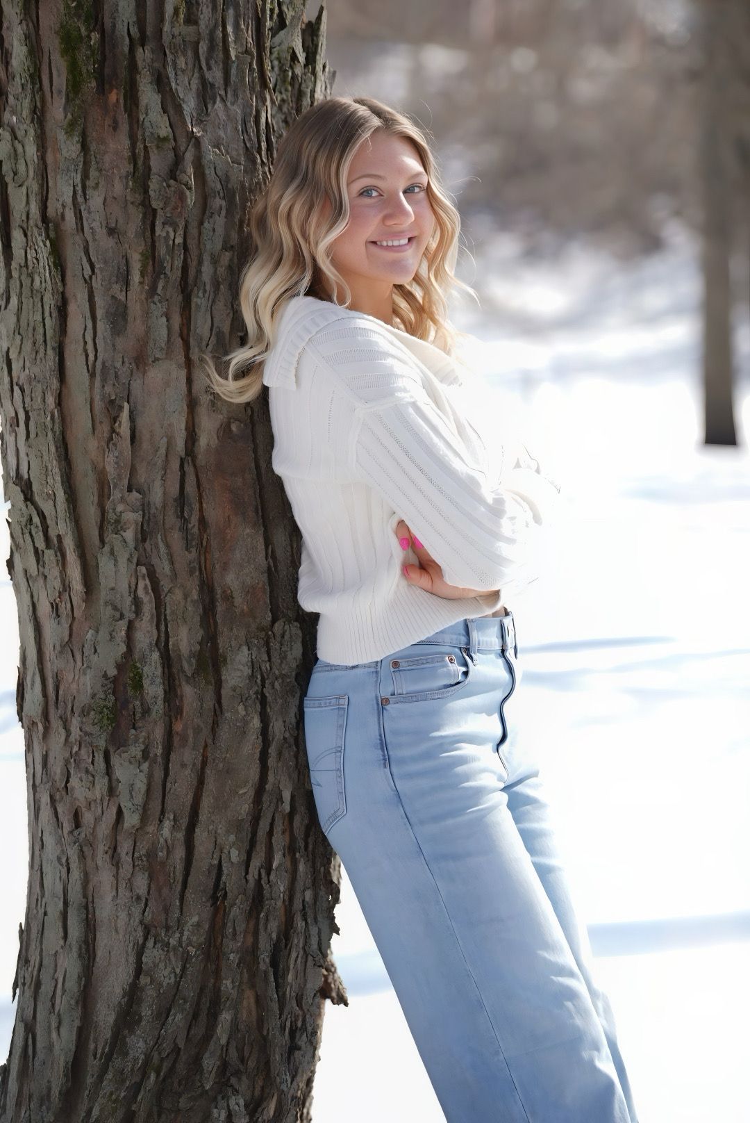 Person in white sweater and light blue jeans poses playfully against a tree trunk in snowy winter setting.