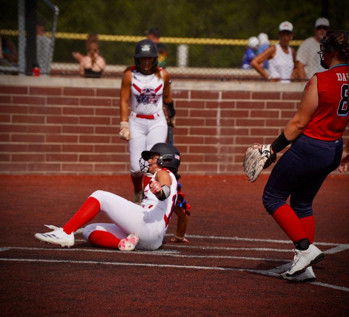 Softball player sliding into home plate during a competitive game on a dirt field.