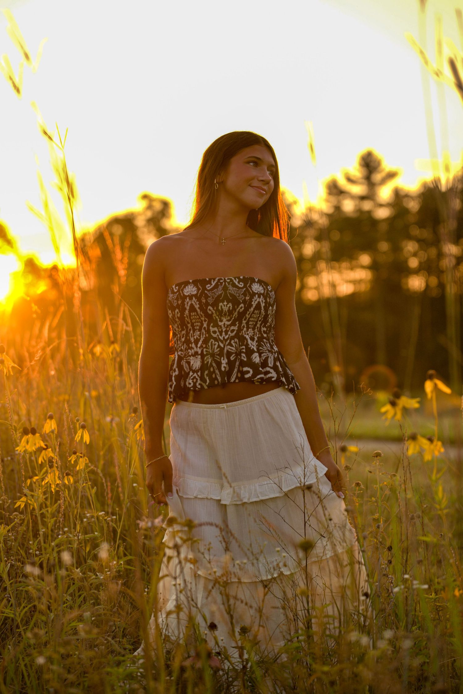 Young woman in strapless top and white tiered skirt standing in golden sunset field with wildflowers.