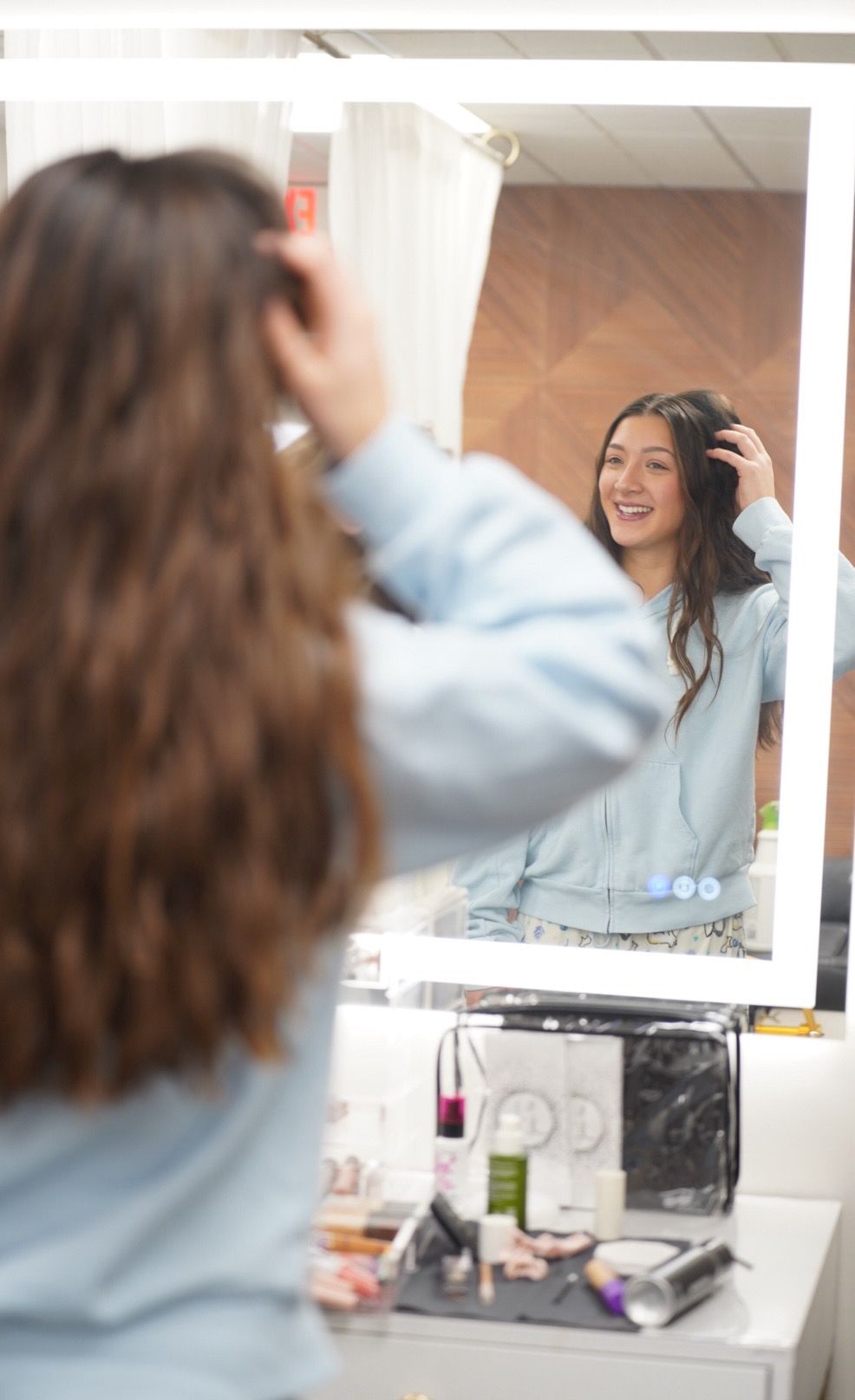 Someone in a light blue sweater looks at their reflection while styling their long dark hair in a bathroom mirror.