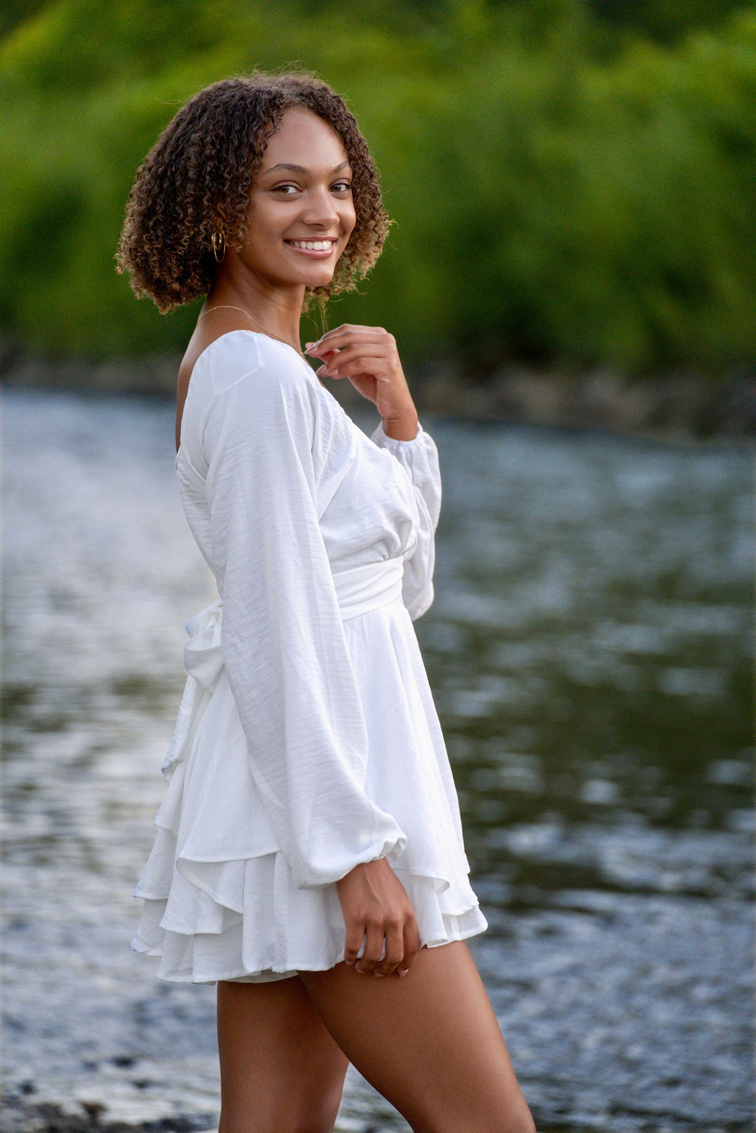 Young woman in white wrap mini dress with ruffles smiling by a river with lush green trees.