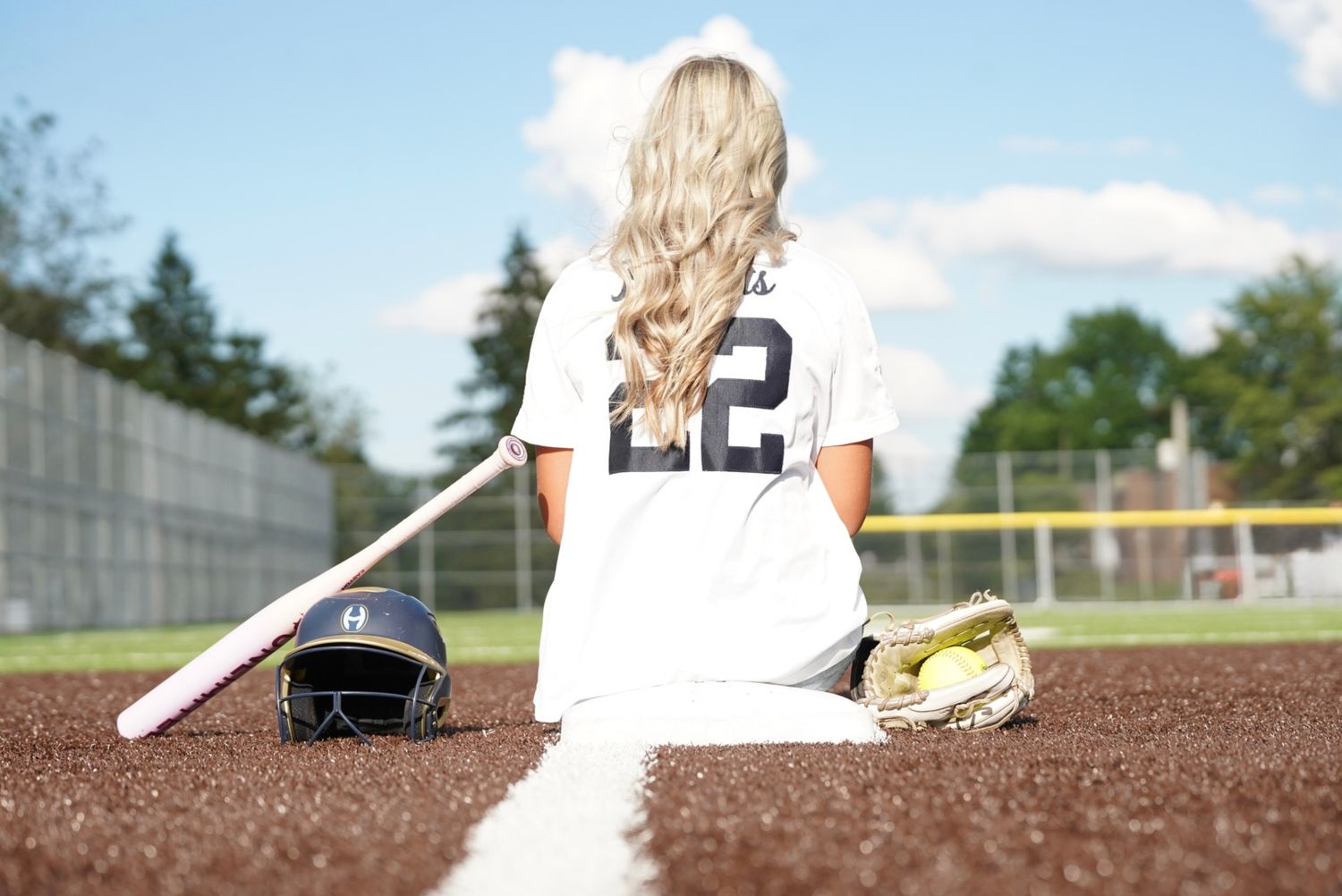 Softball player #22 sitting on field with bat, helmet, and glove on a sunny day.