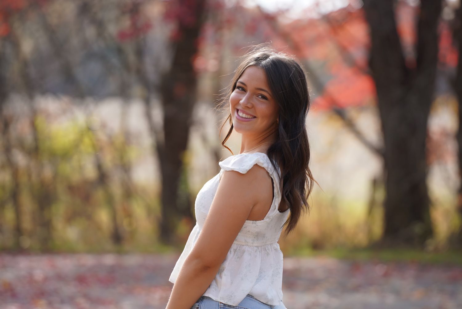 A person in a white tank top and jeans smiles while posing outdoors with autumn foliage in the background.