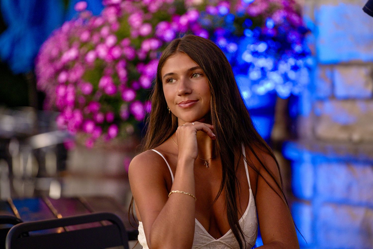 Young woman with long brown hair smiling gracefully, seated outdoors near vibrant pink floral decorations at night.