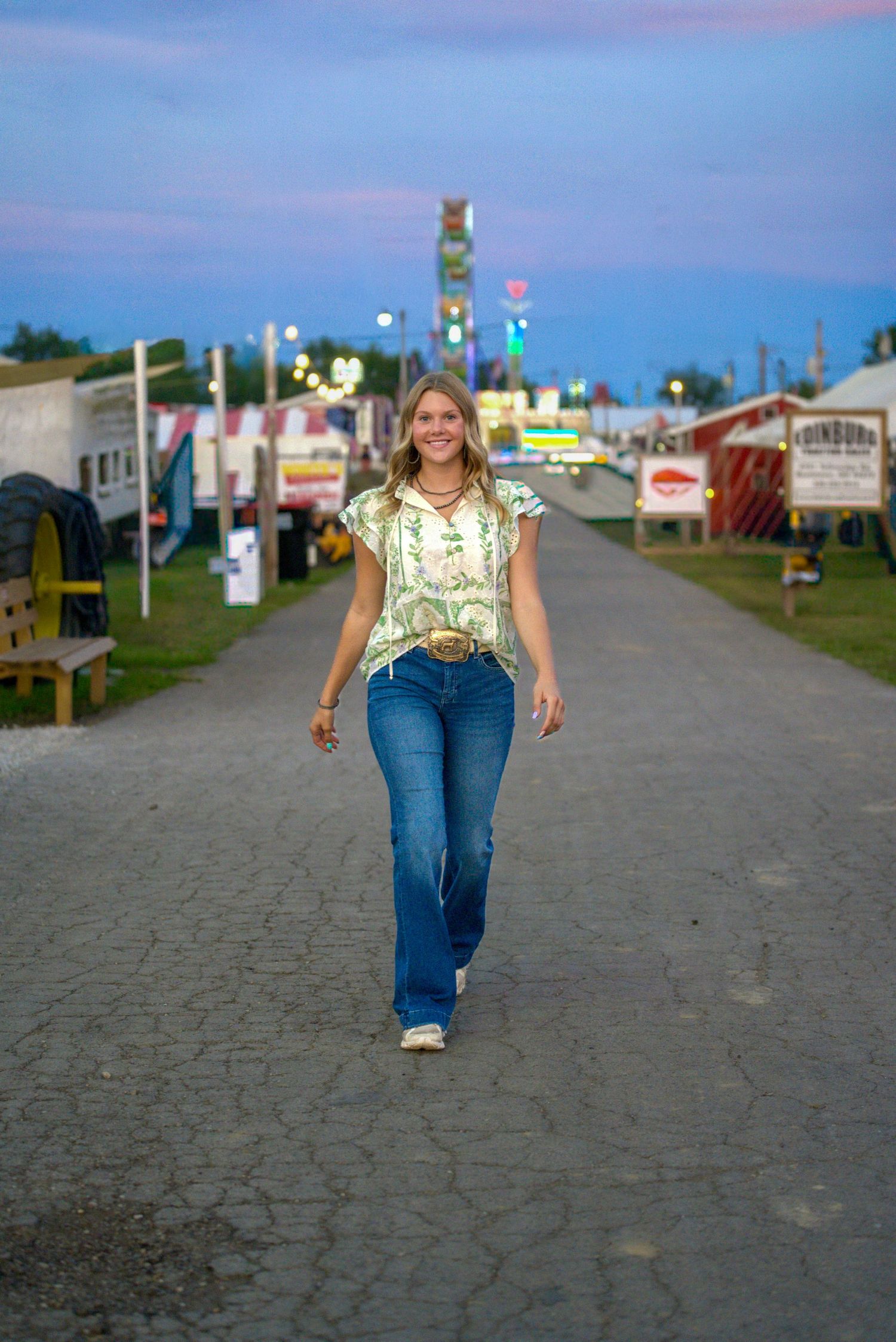 Young woman in floral top and flared jeans walking at a county fair at dusk with colorful lights.