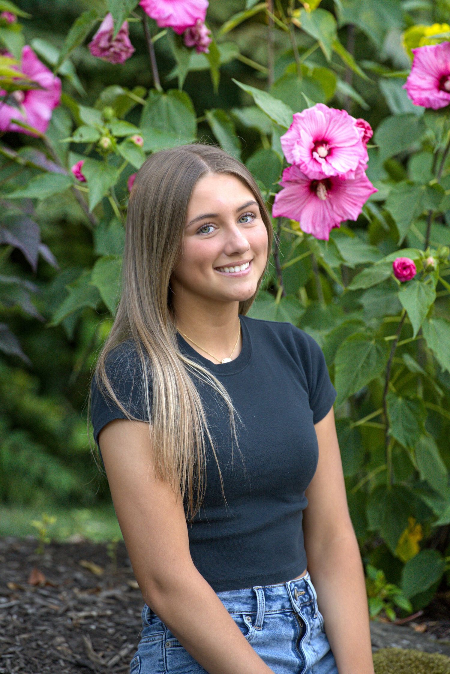 Smiling young woman with long blonde hair poses in front of vibrant pink hibiscus flowers in a lush green garden.