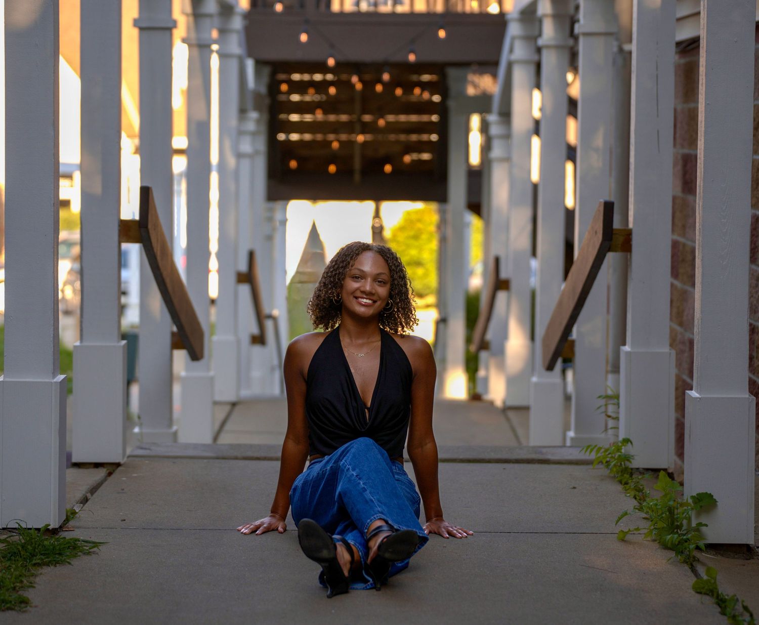 Young woman in black halter top and blue skirt kneeling and smiling in a white pergola walkway.