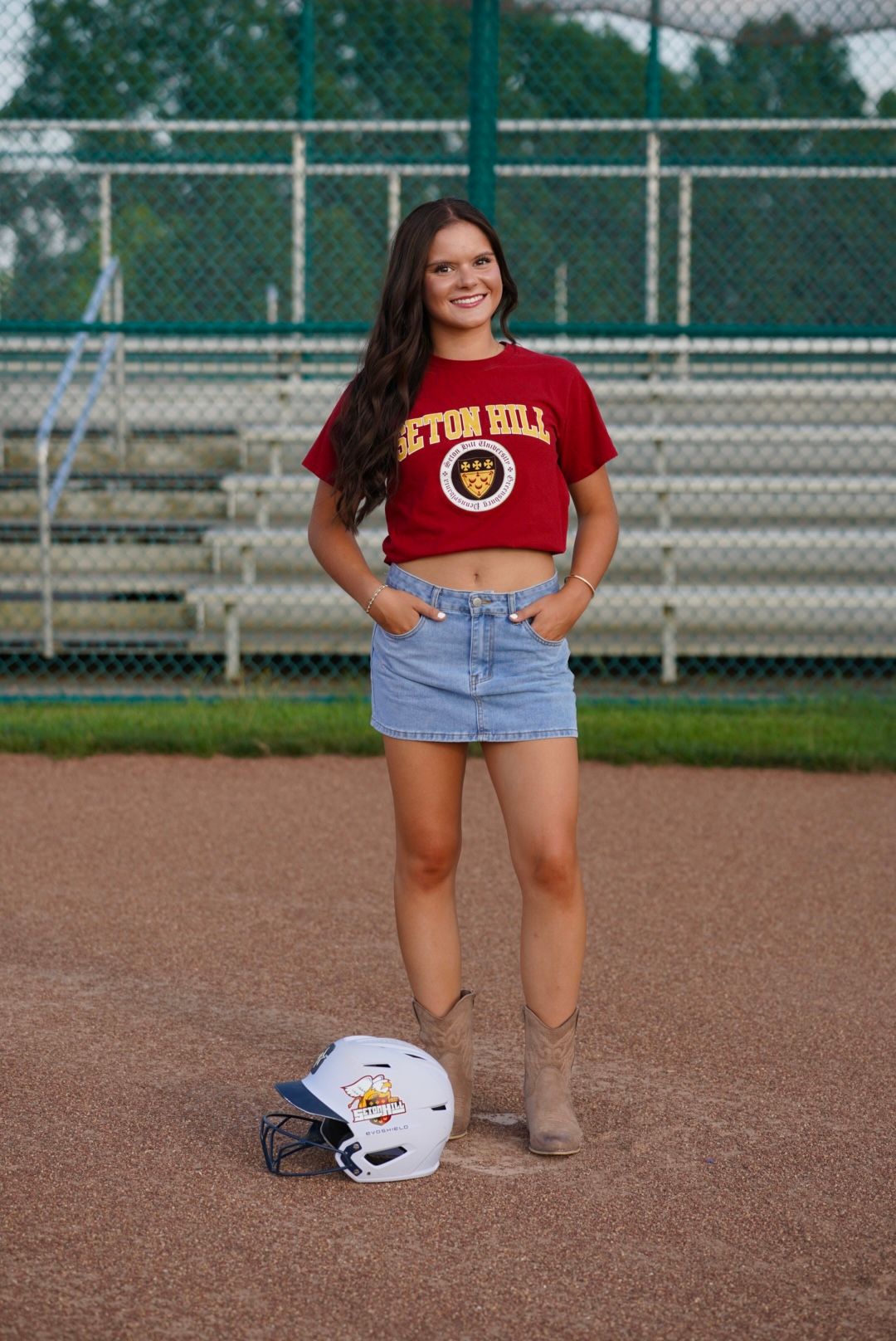 Young woman in Seton Hill crop top and denim skirt poses on baseball diamond with batting helmet at her feet.