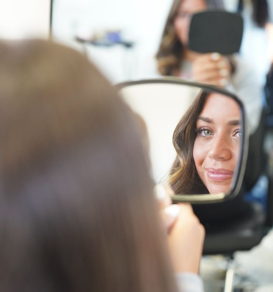 Reflection in handheld mirror shows wavy brown hair and a gentle expression during a beauty session.