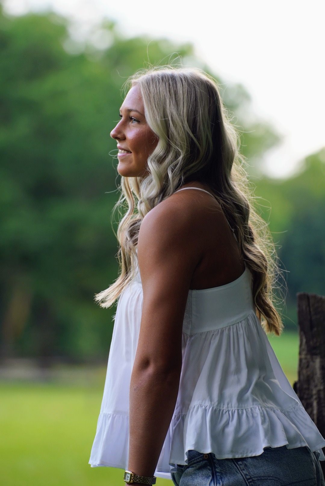 Smiling blonde woman in white sleeveless top and jeans standing outdoors with green trees in background.