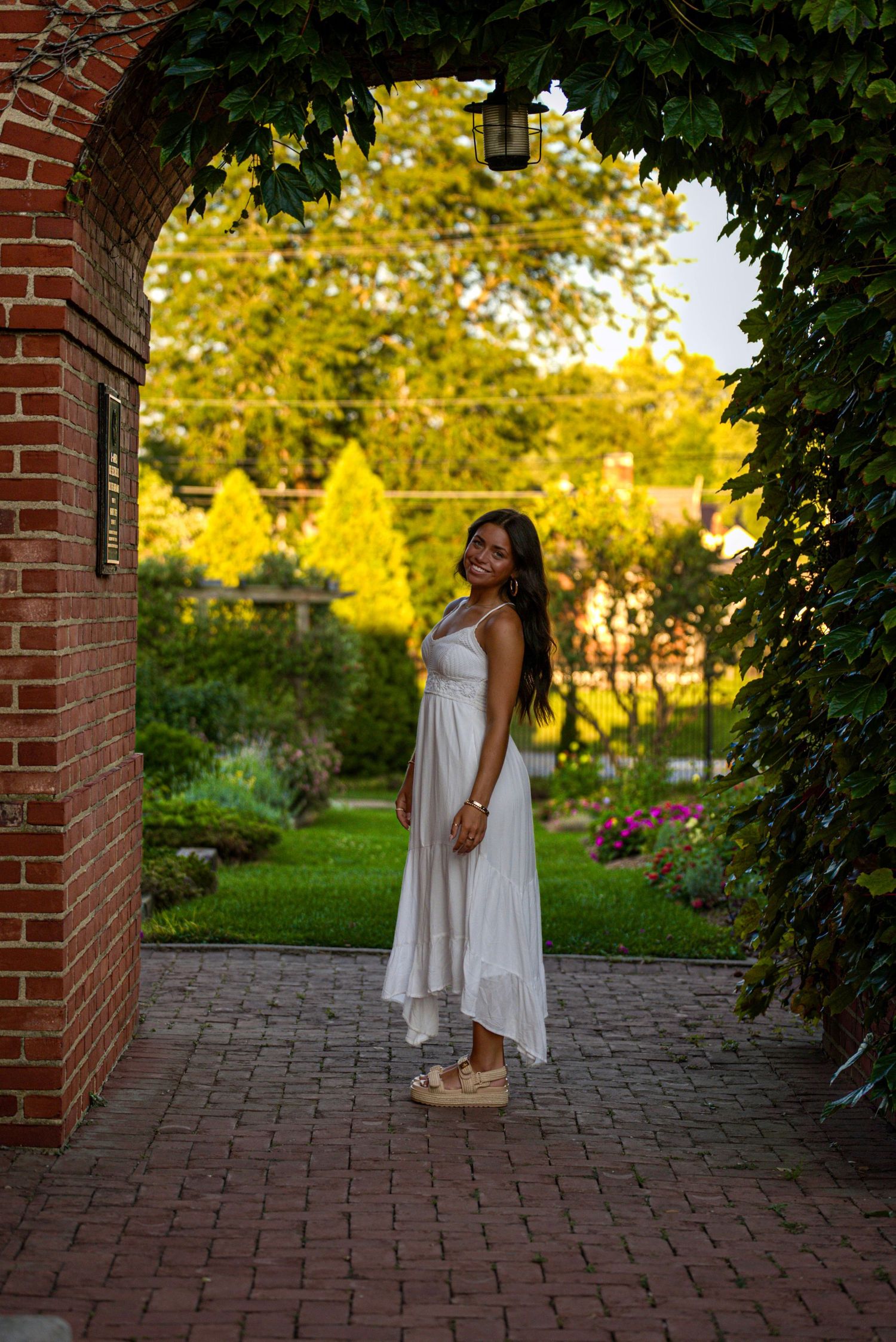 Woman in white dress posing under ivy-covered brick arch in lush garden at golden hour.