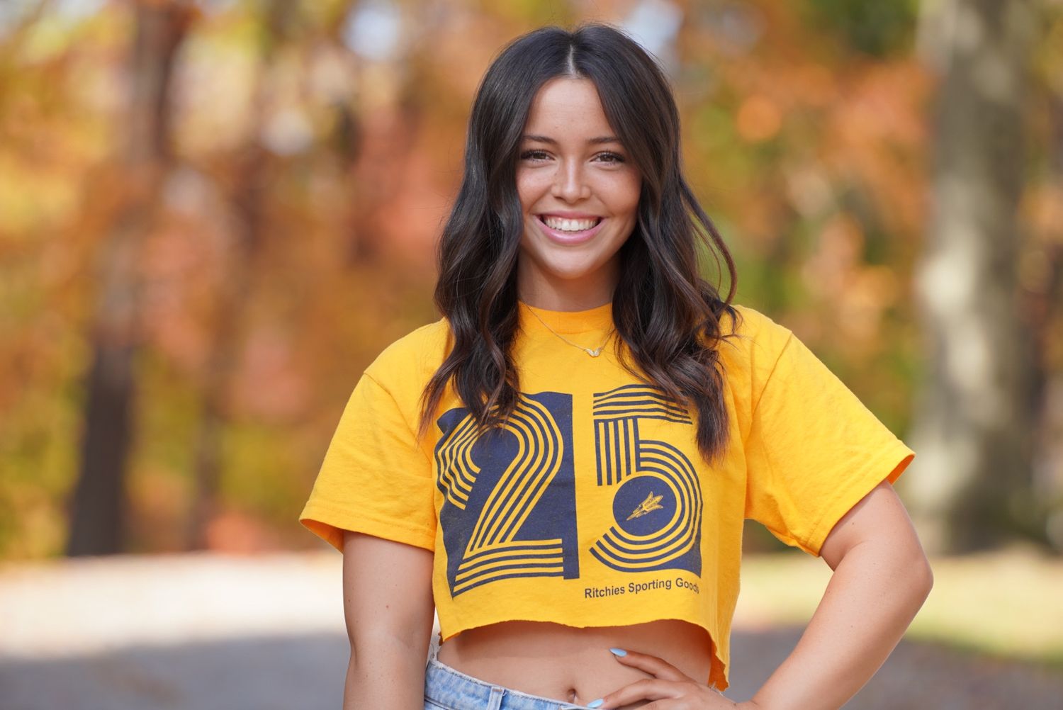 Person wearing a bright yellow crop top with number 25 smiles outdoors against autumn foliage background.