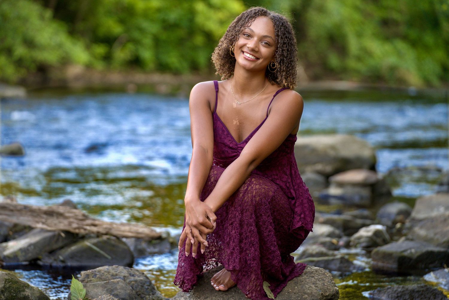Young woman in burgundy lace dress sitting on rocks beside a serene river, smiling warmly.