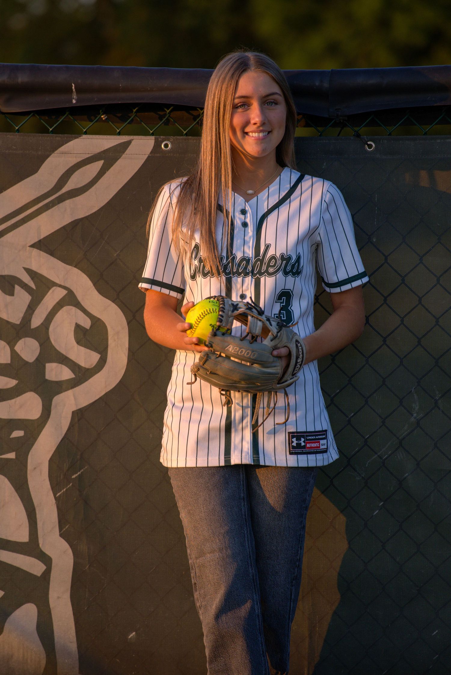 Young softball player in Crusaders pinstripe jersey holding glove and ball, posing outdoors in golden hour light.