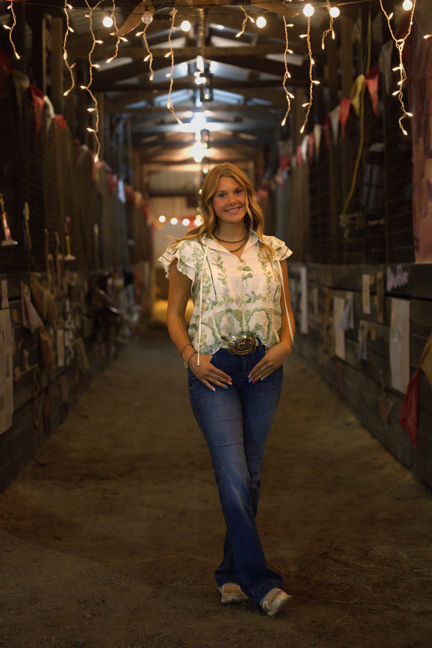 Woman in floral top and flared jeans poses in a rustic barn hallway decorated with string lights and colorful bunting.
