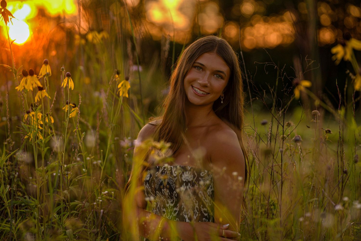 Young woman smiling in a wildflower field during golden hour sunset, wearing a floral top.