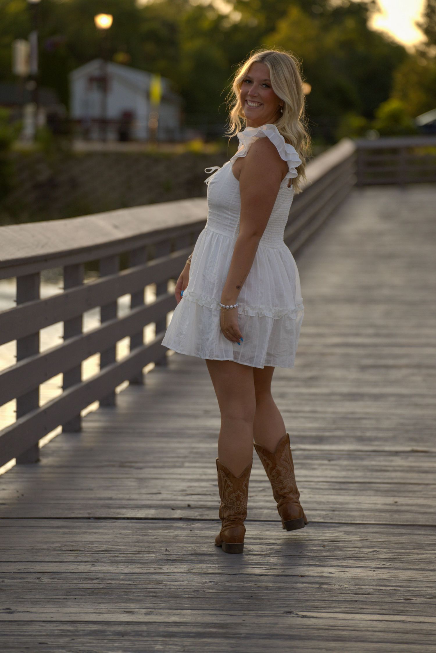 Smiling blonde woman in white dress and cowboy boots posing on wooden bridge at golden hour.
