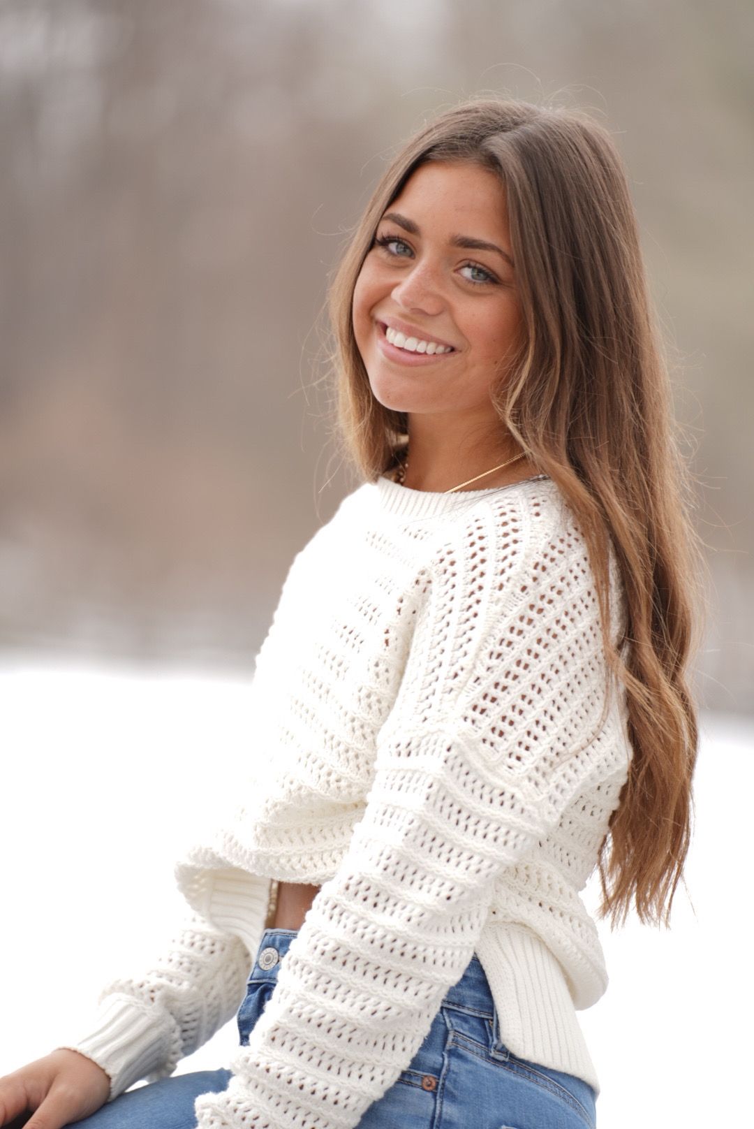 A person in a cozy white knit sweater and jeans poses playfully against a snowy winter backdrop.