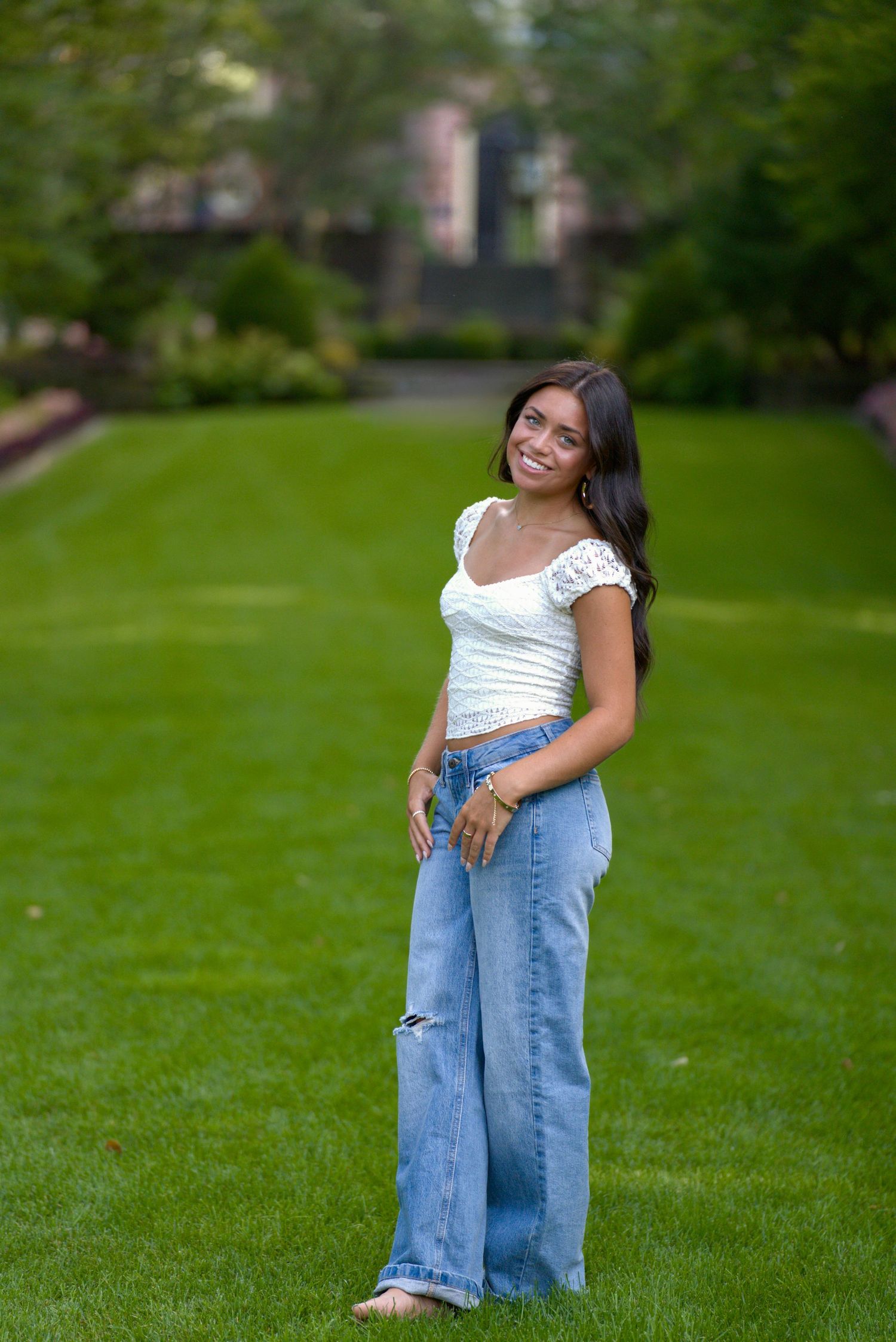 Young woman smiling in white crop top and wide-leg jeans standing on green lawn in outdoor park setting.