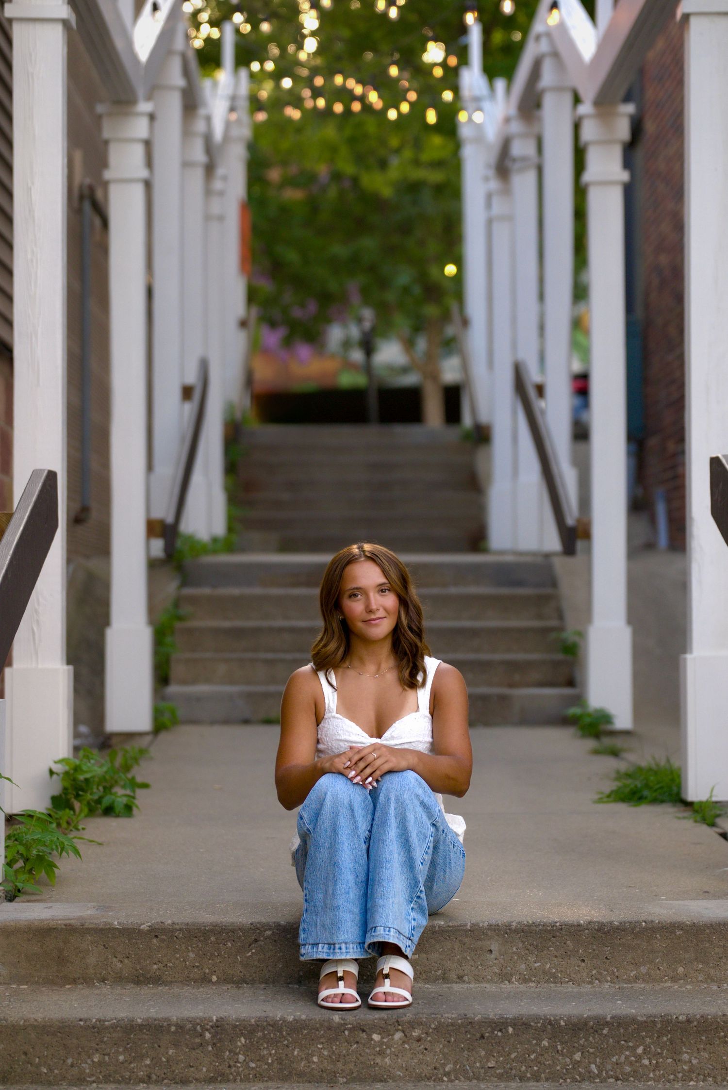 Young woman in white top and jeans sits on outdoor steps with white columns and string lights above.