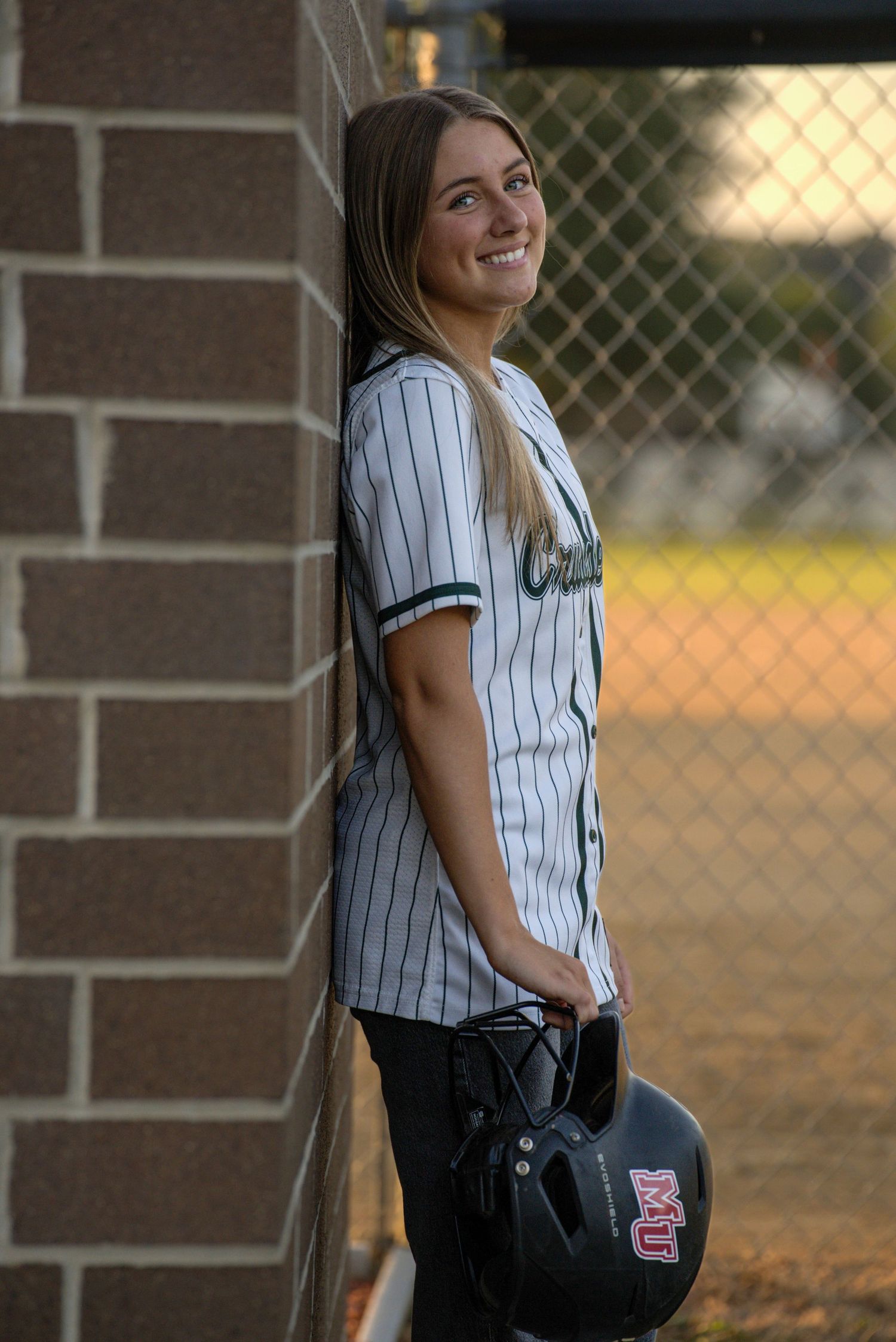 Young female softball player in pinstripe jersey leaning against brick wall holding batting helmet at baseball field.