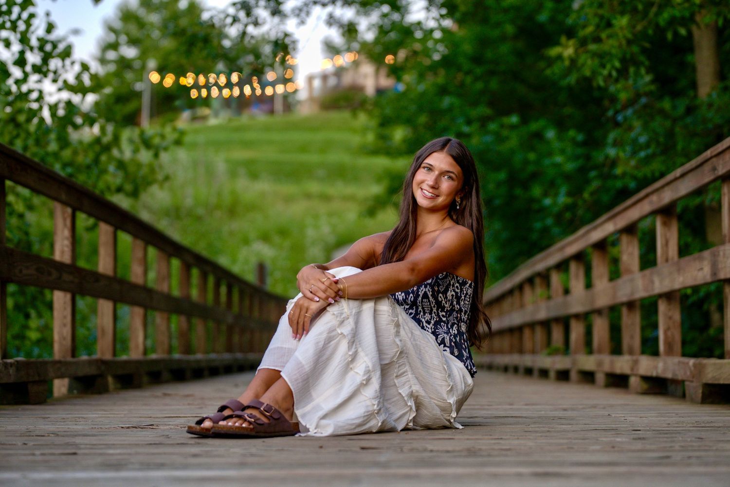 Young woman with long dark hair sits smiling on a wooden bridge, wearing a floral top and white skirt with sandals.