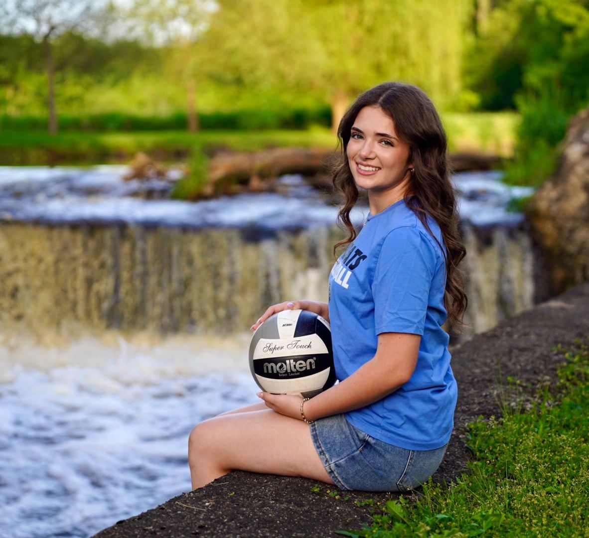 Young woman in blue shirt holding a Molten volleyball, posing by a scenic waterfall outdoors.