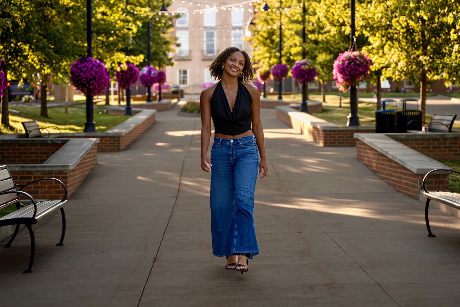 Young woman in black halter top and wide-leg jeans walking on a sunny campus pathway.