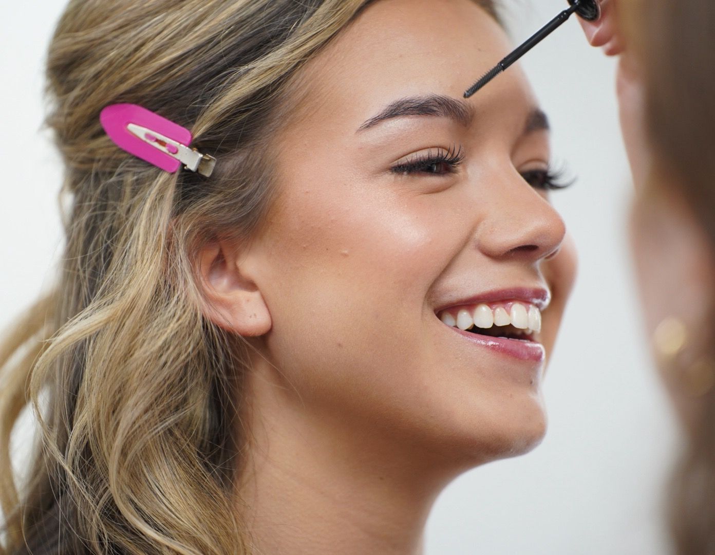Person smiling while having makeup applied with a pink hair clip visible in their blonde hair.
