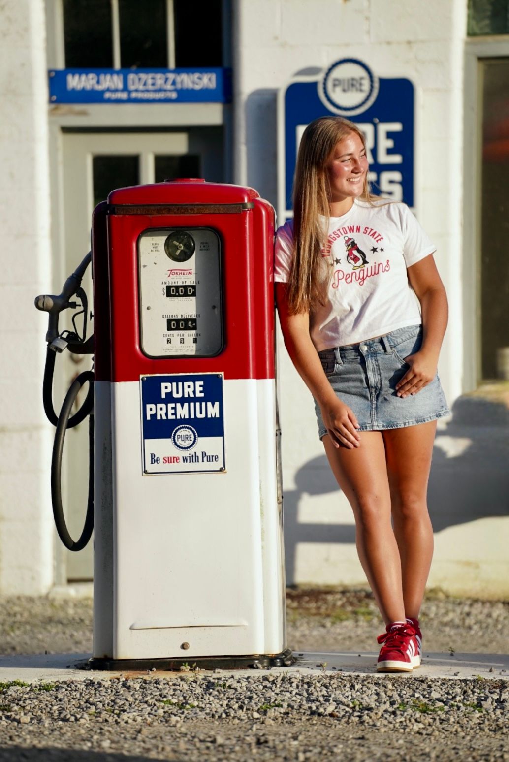 Young woman in denim skirt poses beside vintage Pure Premium gas pump at retro gas station.