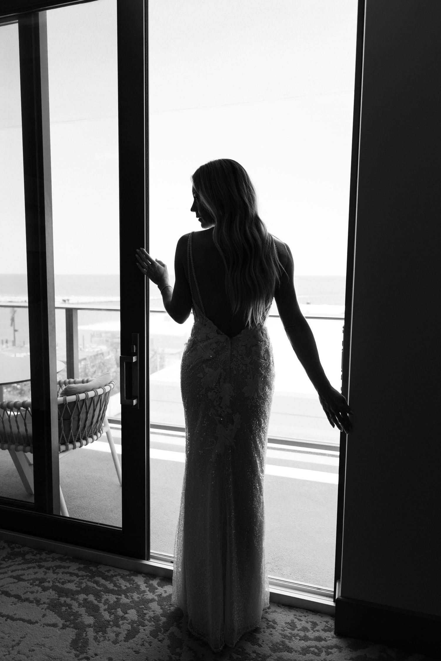 Black and white silhouette of a bride in a backless lace gown standing by ocean-view doors.