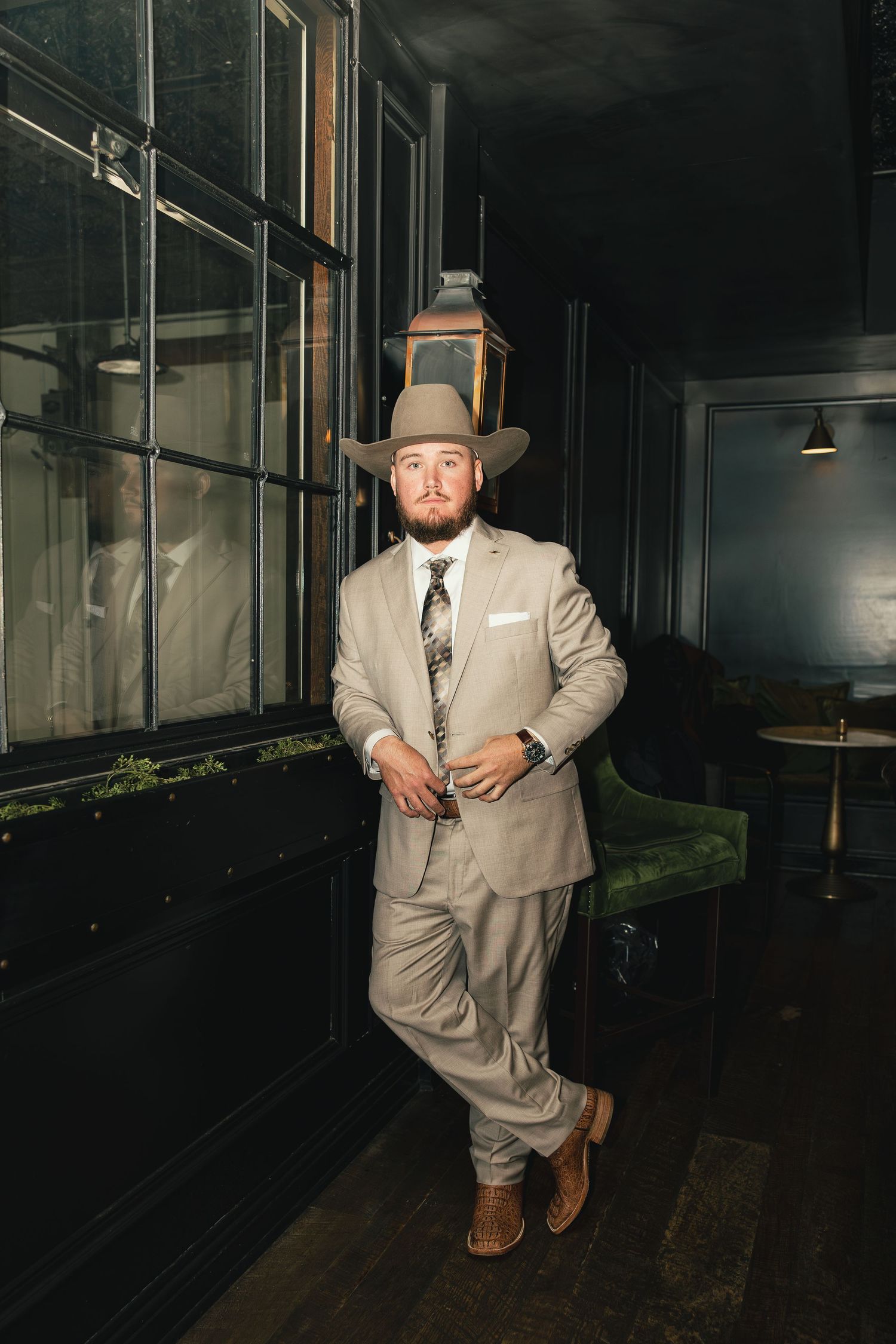 Man in beige suit and cowboy hat poses confidently in a dimly lit upscale bar interior.