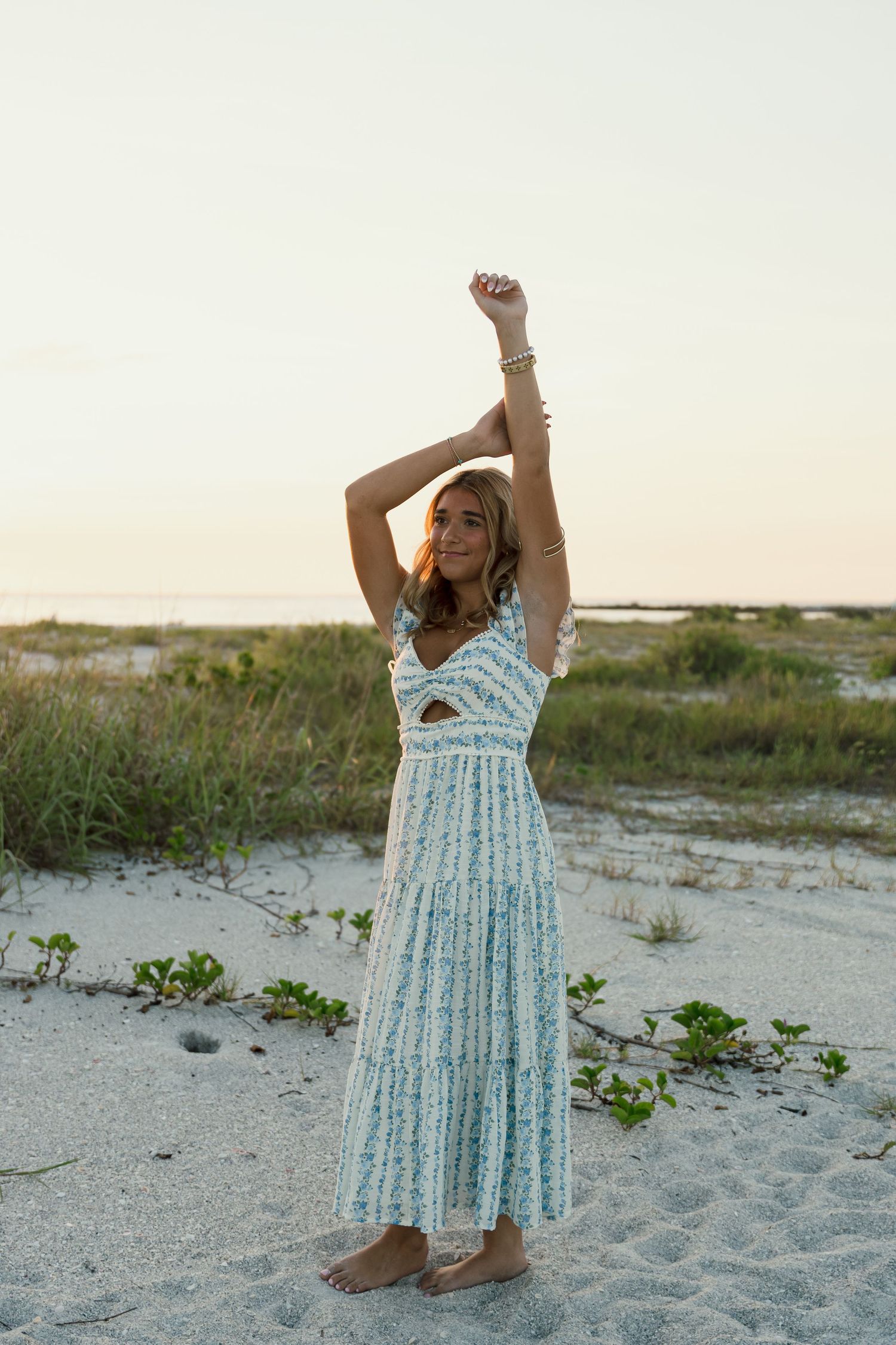 Woman in blue floral maxi dress poses on sandy beach at sunset with arms raised.