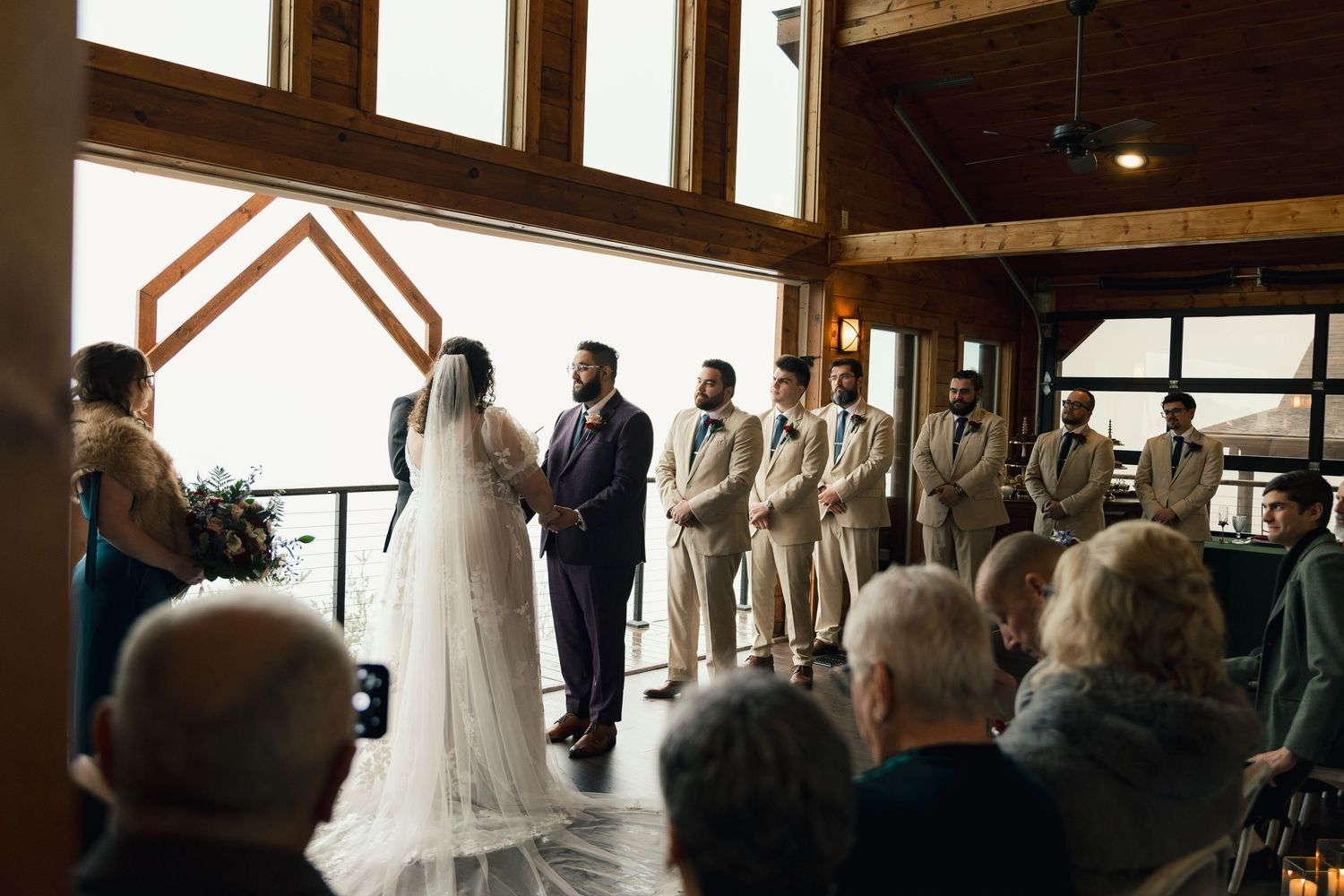 Bride and groom exchange vows at indoor wedding ceremony with wooden arch and groomsmen.