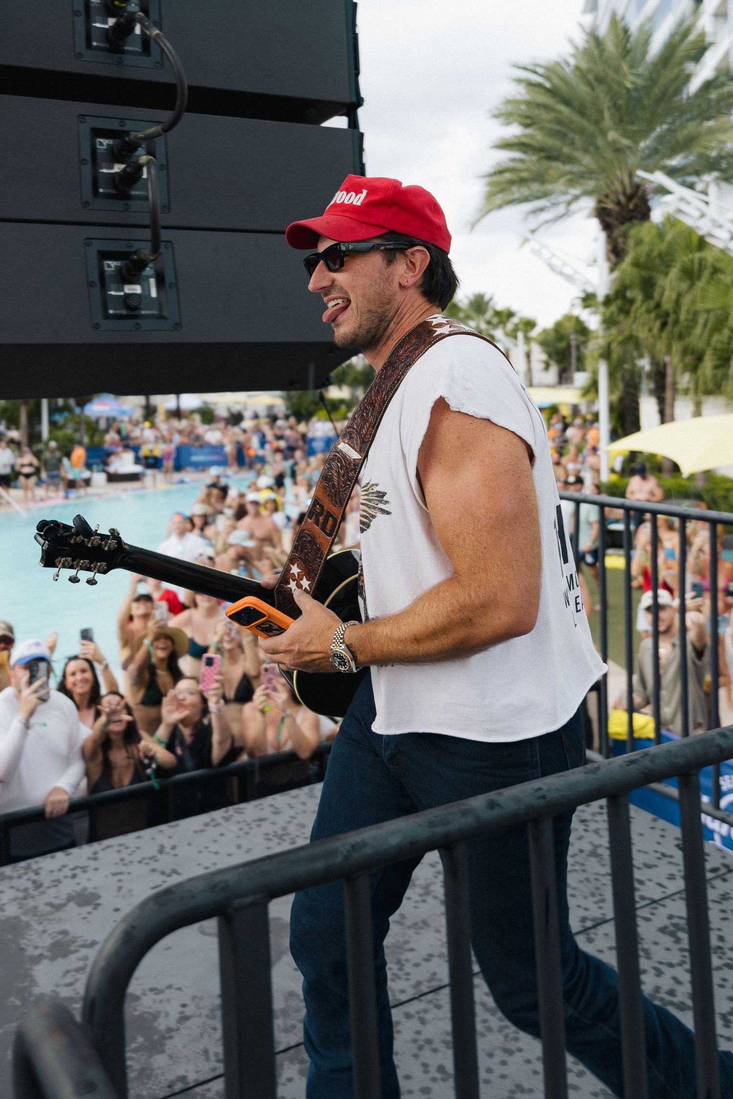 Male guitarist in red cap and white sleeveless shirt performs on outdoor stage near pool for large crowd.