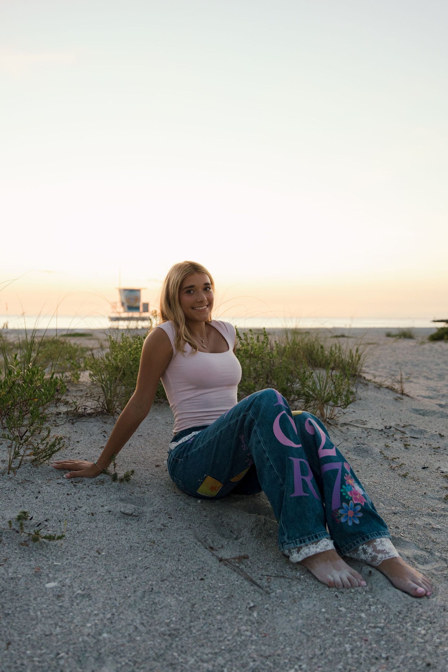 Blonde woman sitting on sandy beach at sunset wearing jeans and white tank top with lifeguard tower in background.