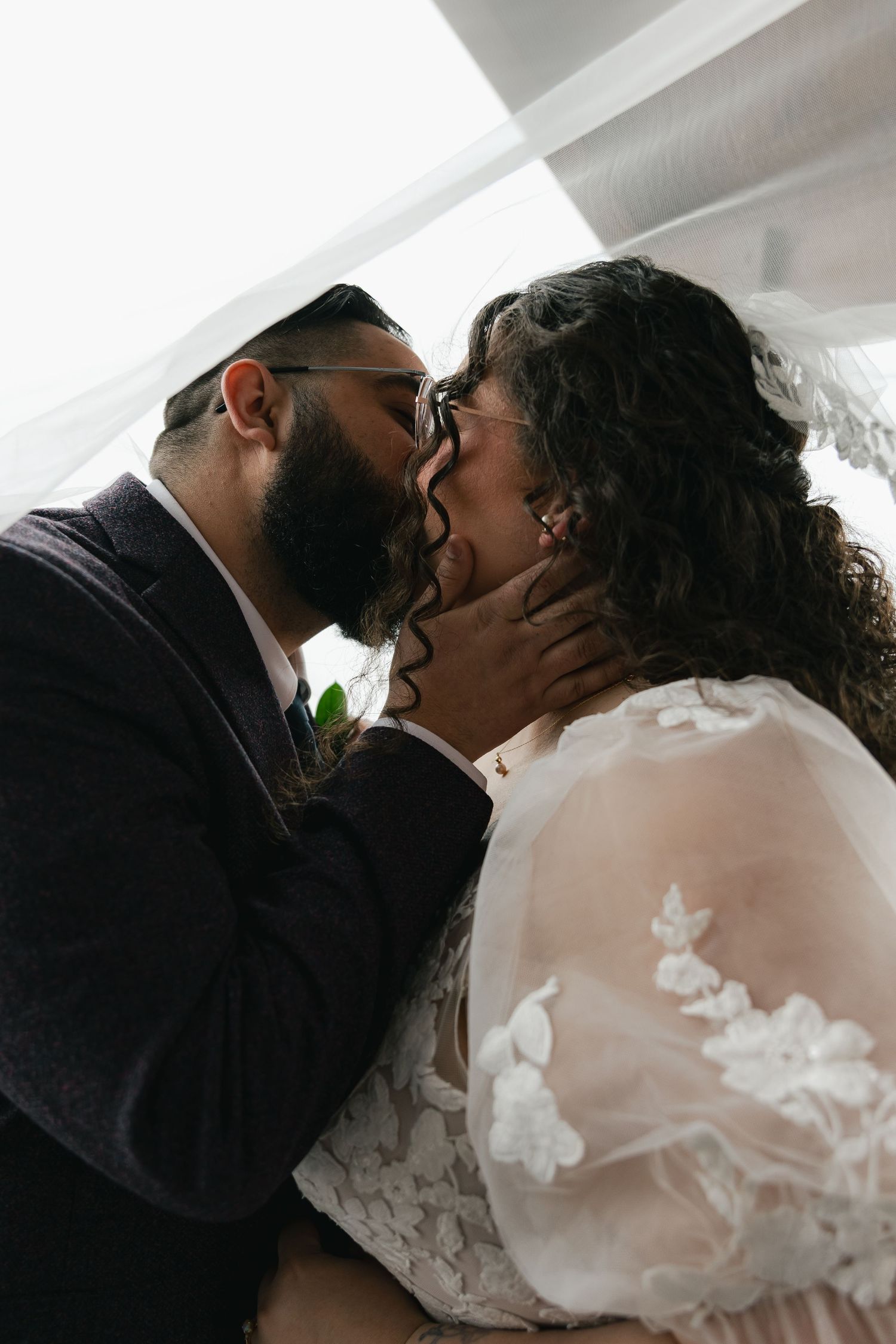 Bearded groom tenderly kisses bride in lace dress under wedding canopy.