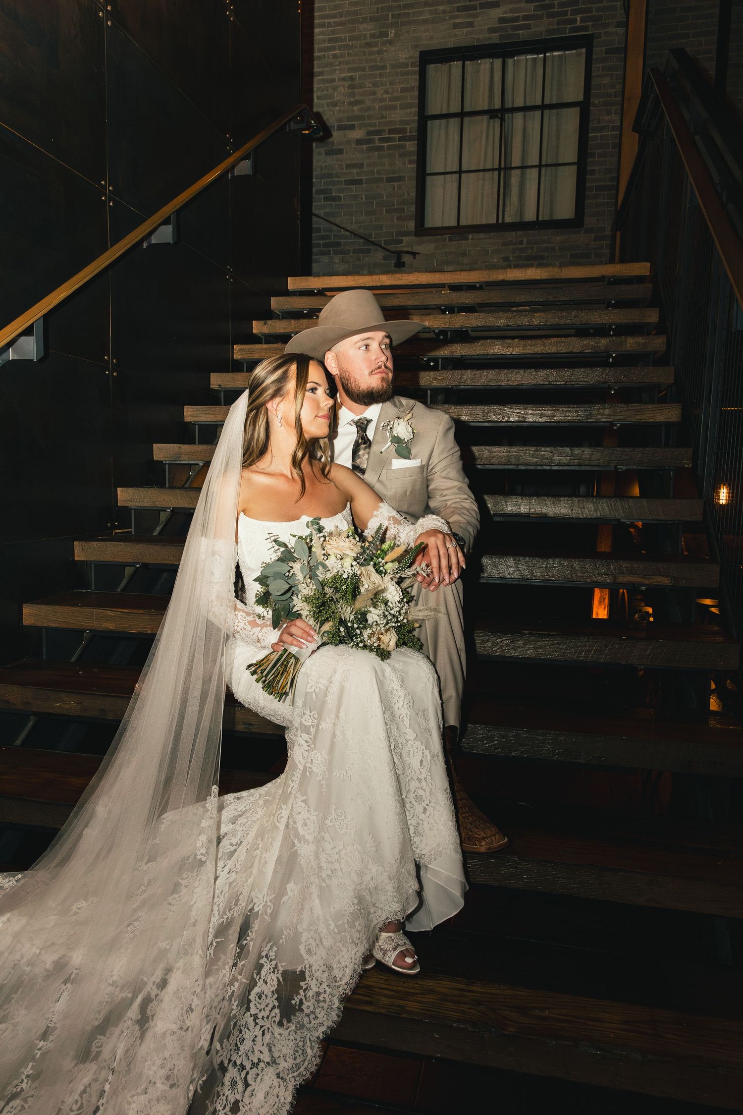 Bride and groom posing on wooden stairs, groom wearing a tan suit and cowboy hat.