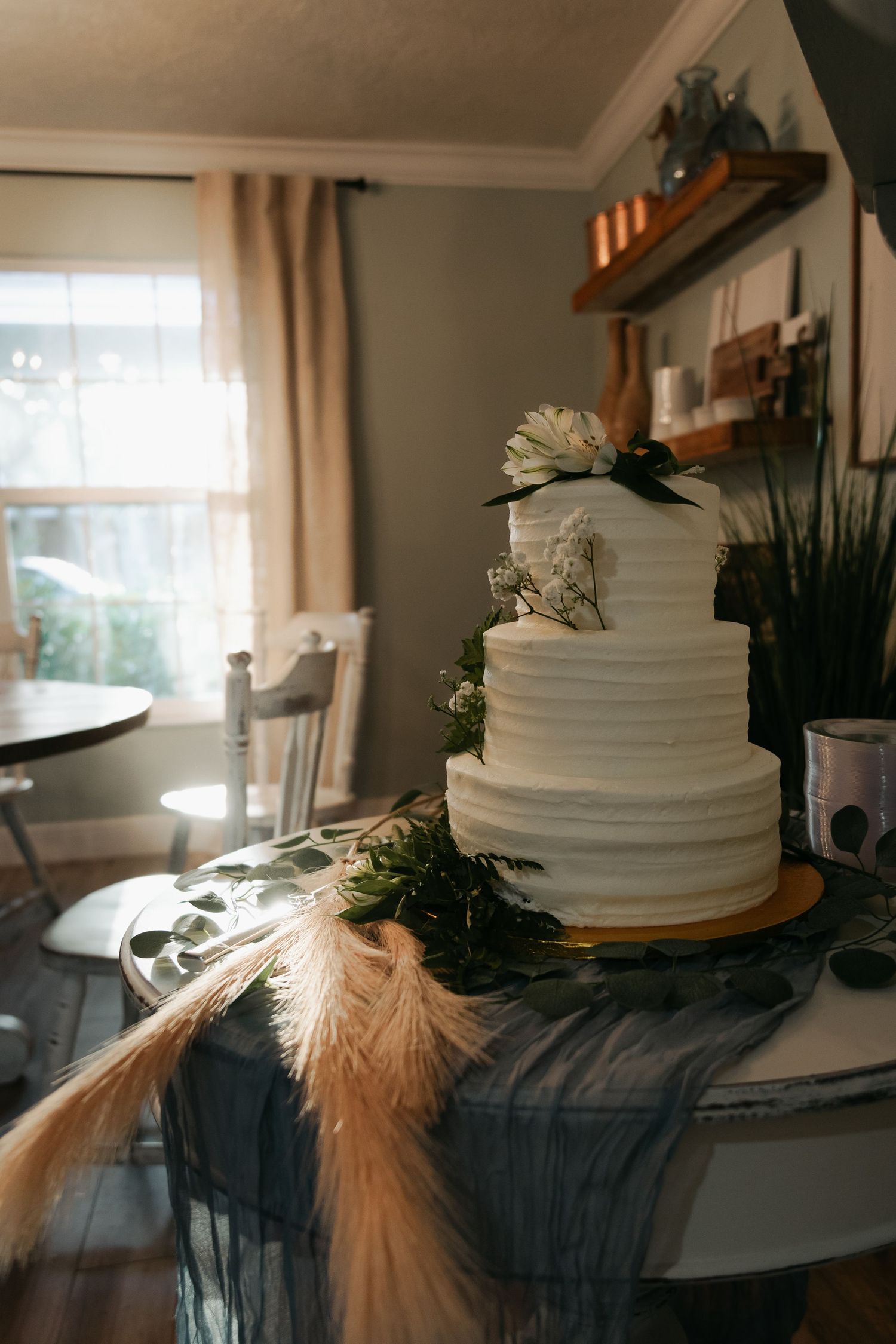 Elegant three-tier white wedding cake adorned with flowers and pampas grass on a gold stand.
