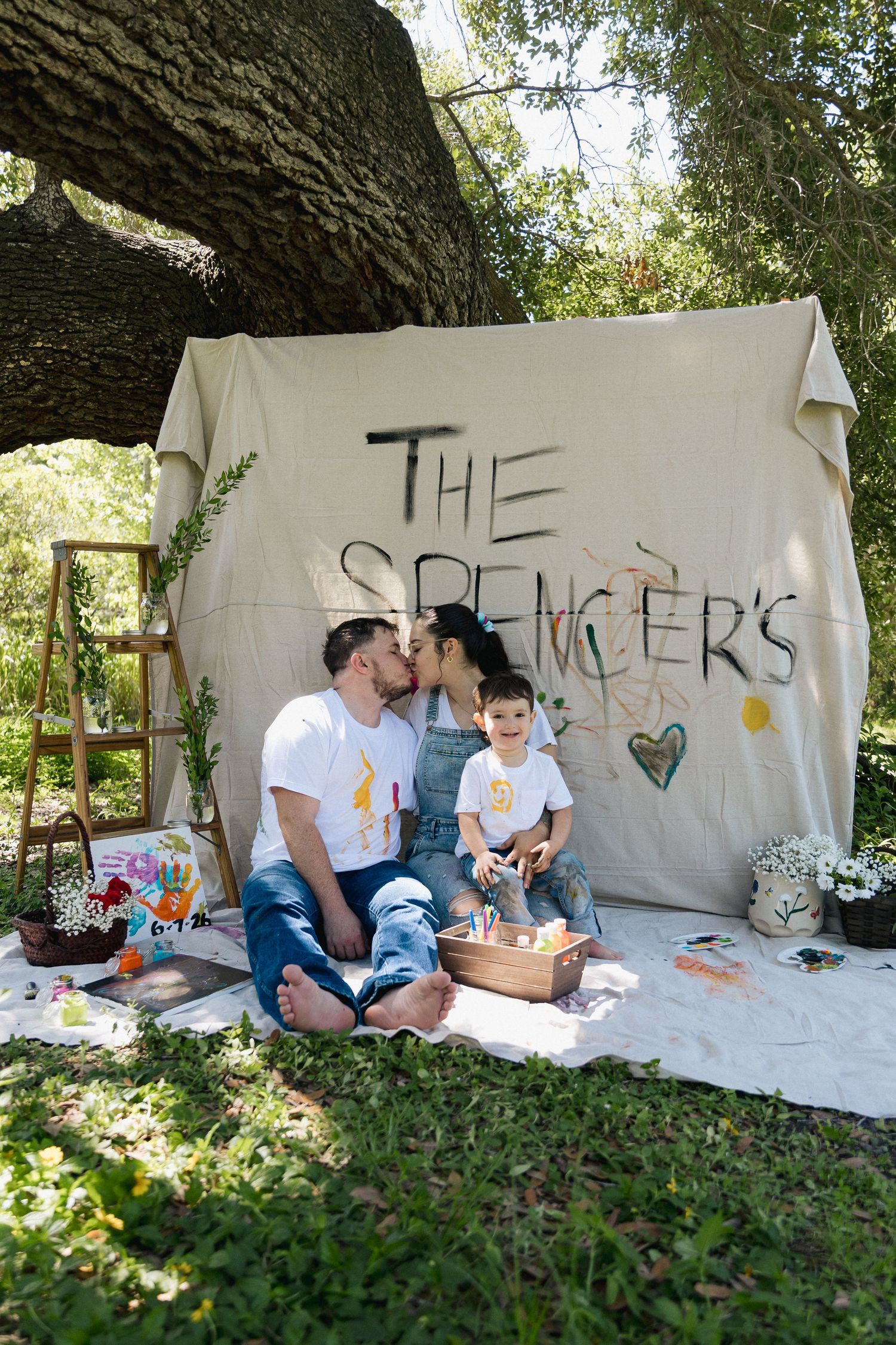 The Spencer family sits together outdoors under a tree, kissing and smiling in front of a painted canvas backdrop.