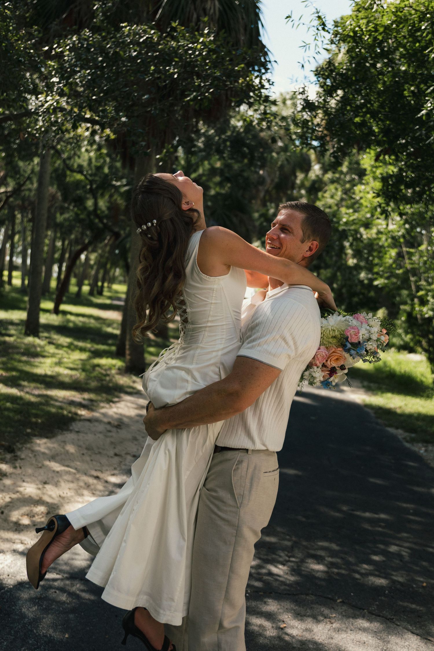 Joyful groom lifting laughing bride in white dress while holding bouquet on tree-lined path.