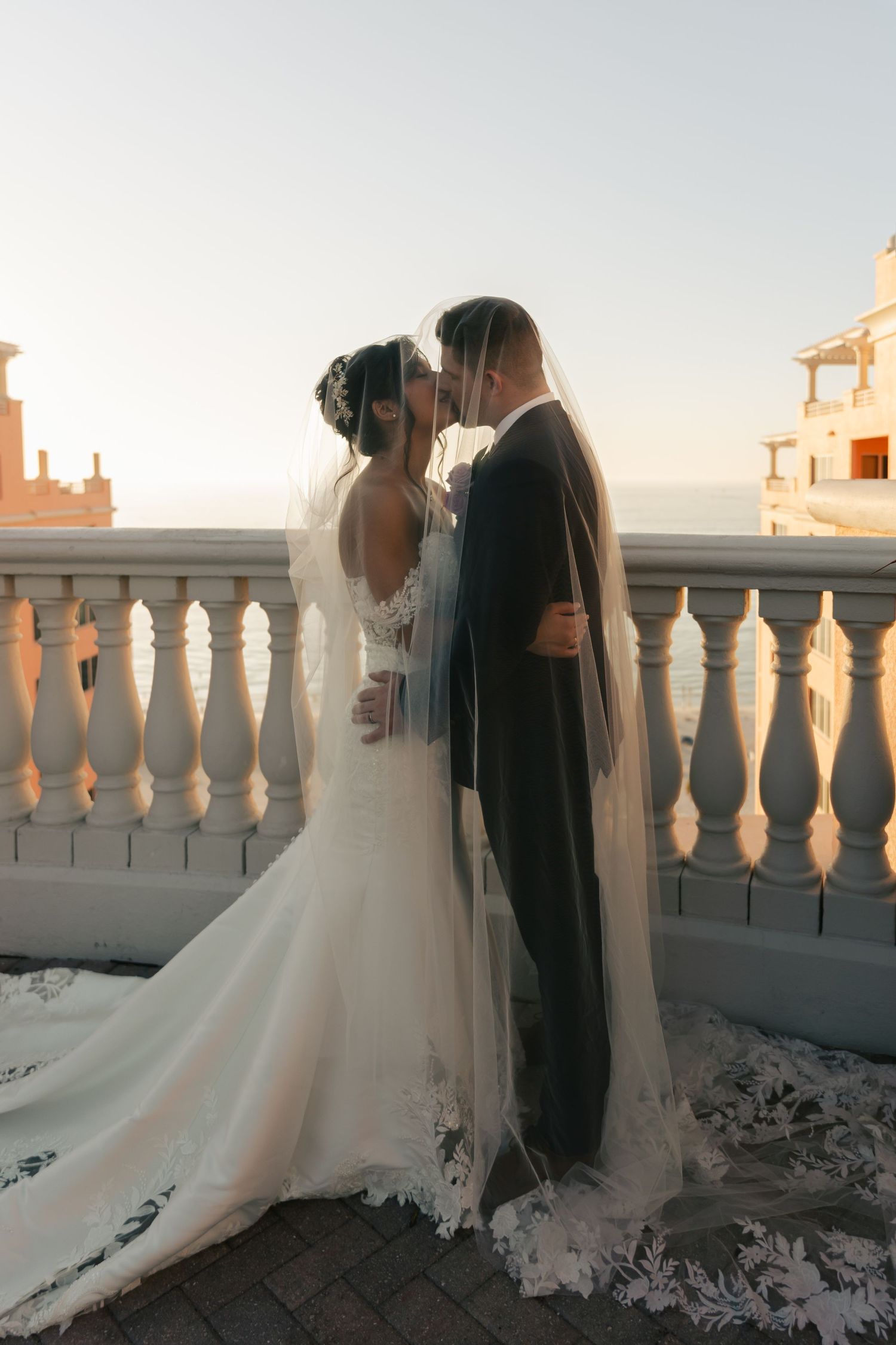 Bride and groom sharing a romantic kiss beneath a flowing veil on a sunlit coastal balcony.