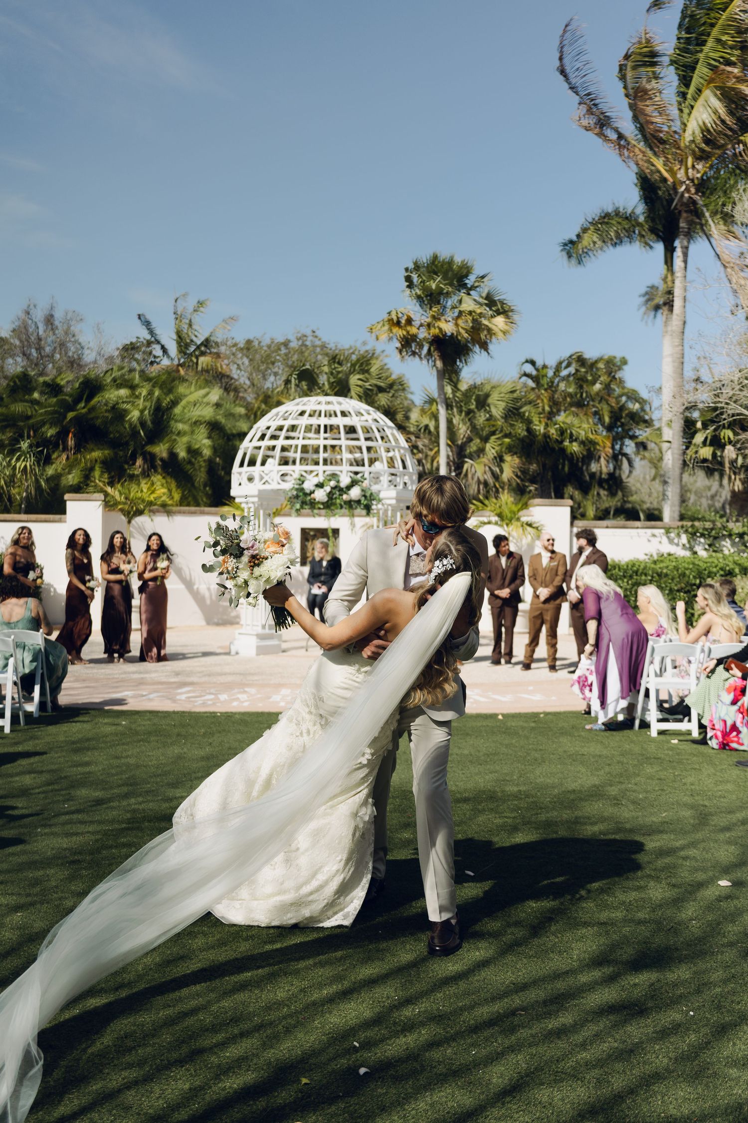 Groom lifts bride in white gown at outdoor wedding ceremony with palm trees.