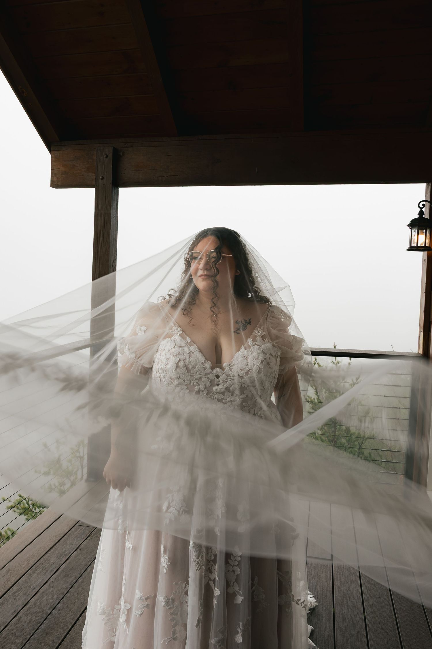 Bride in lace gown with flowing veil billowing on misty outdoor porch.