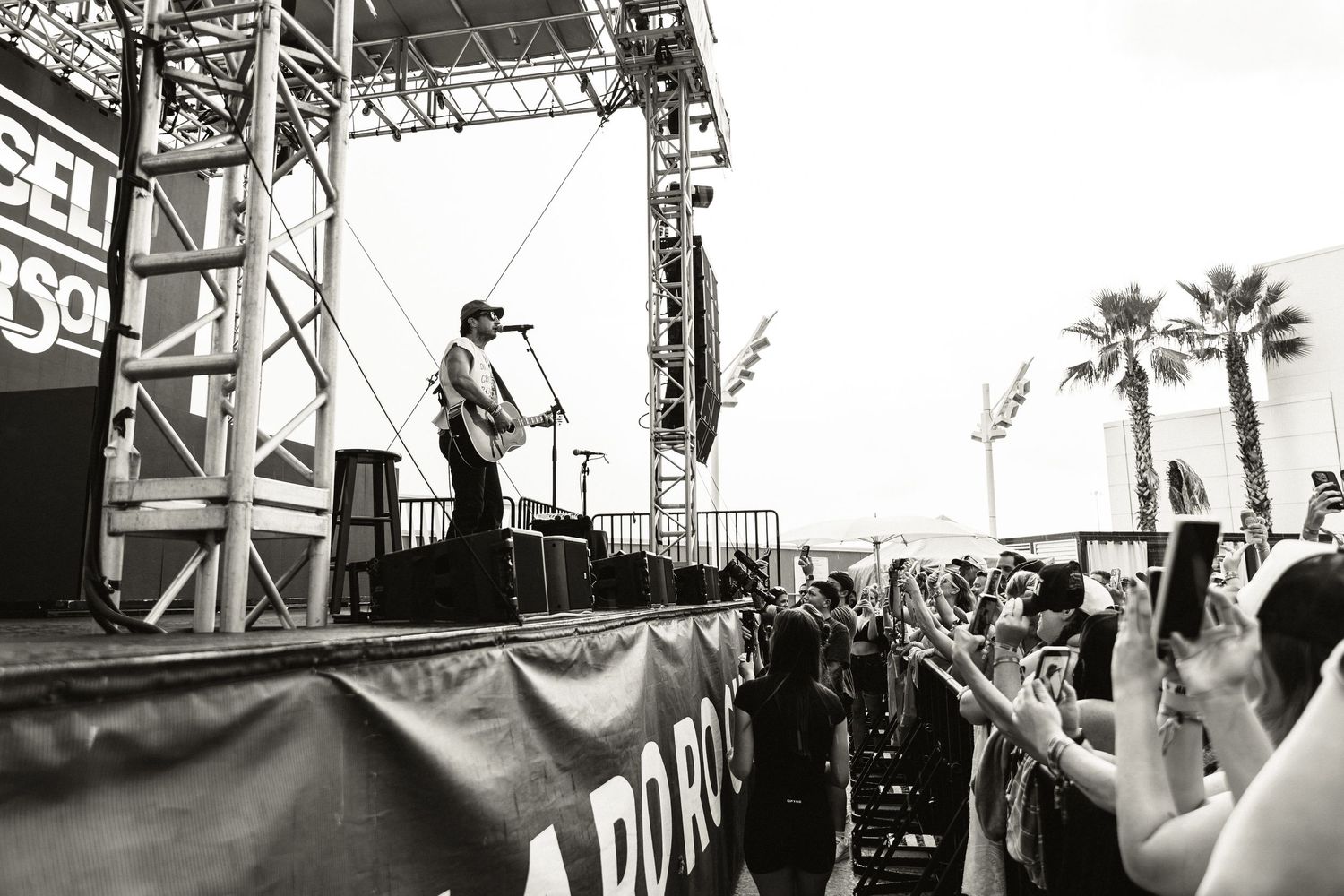 Black and white photo of a guitarist performing on stage at an outdoor music festival with a large crowd.