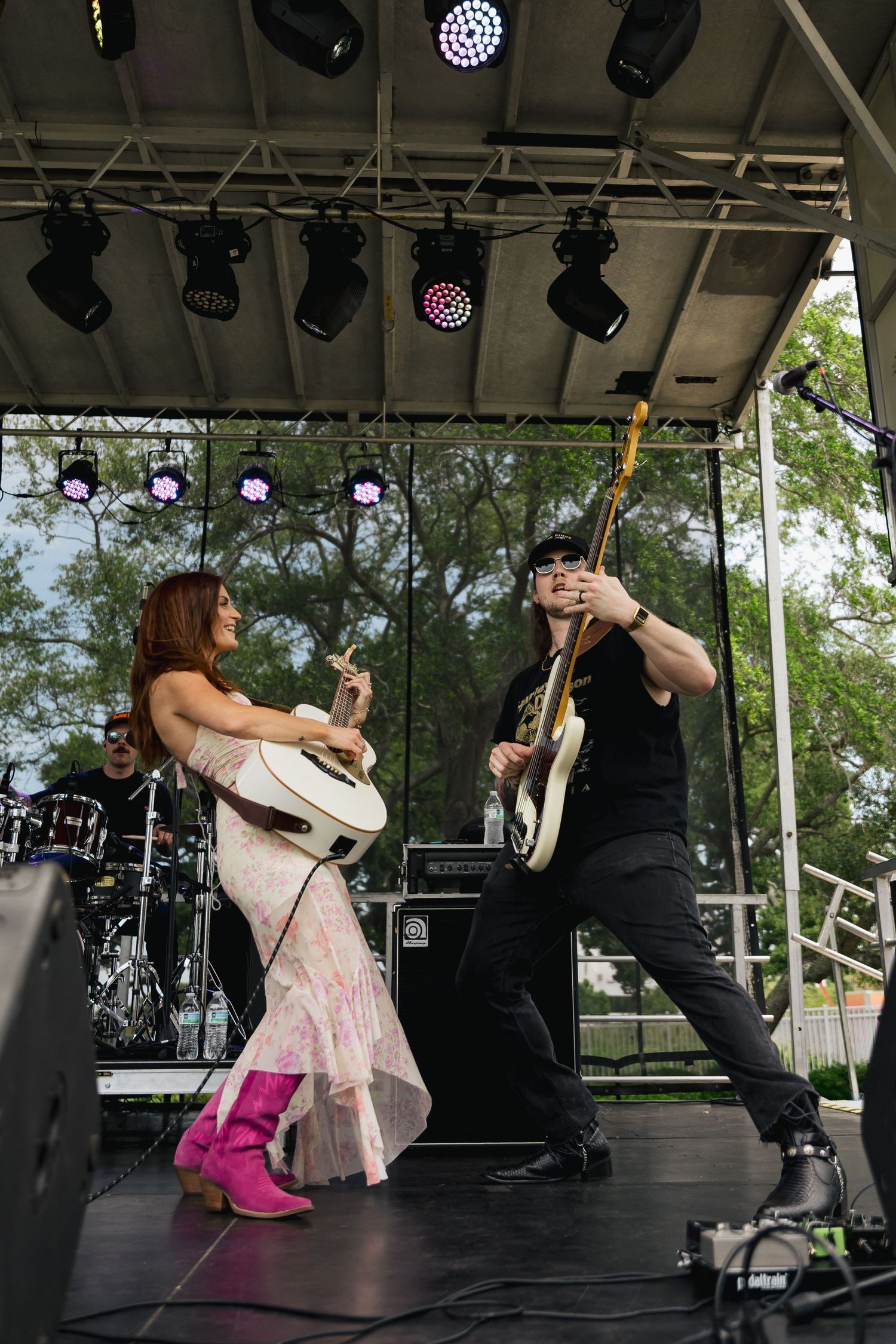 Female and male musicians performing on outdoor stage, woman playing guitar in floral dress with pink boots.