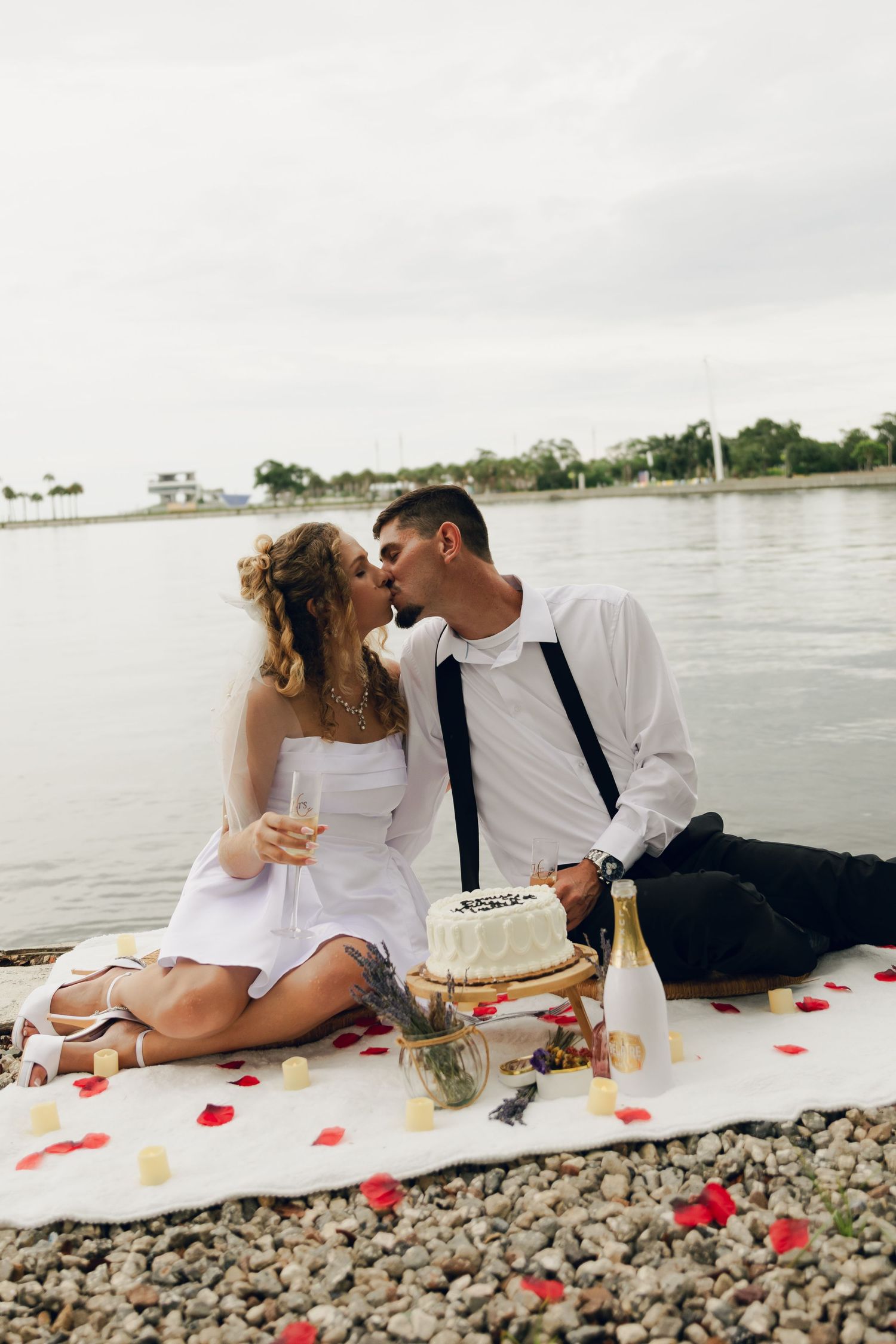 Newlyweds sharing a romantic kiss during a beachside picnic with wedding cake and champagne.