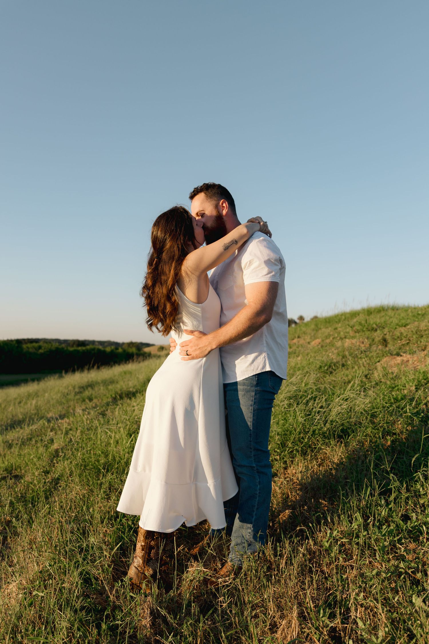 A couple embracing and kissing in a green field at golden hour, woman in white dress, man in jeans and white shirt.