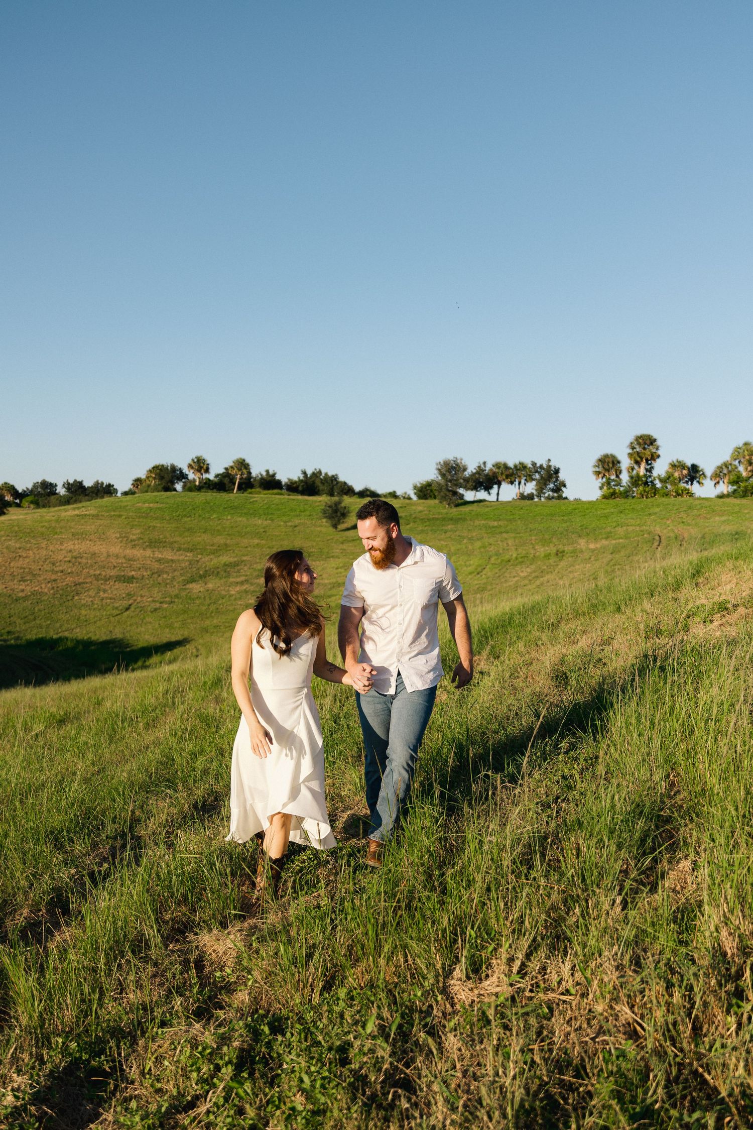 A couple walks hand-in-hand through a lush green field under a clear blue sky, smiling at each other.